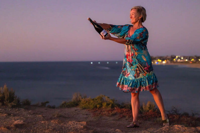 Susan the Champagne Goddess smiling and standing on the limestone cliffs near Port Noarlunga South Australia holding a bottle of champagne about to saber it open.